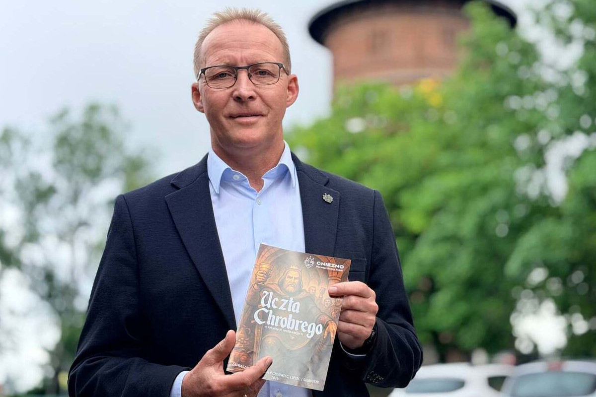 Osoba ubrana w ciemną marynarkę i jasną koszulę stoi na zewnątrz i trzyma w dłoniach folder promocyjny projektu „Uczta Chrobrego”. Na okładce folderu widoczny jest tytuł wydarzenia oraz stylizowana grafika nawiązująca do historycznej tematyki. W tle znajdują się drzewa oraz rozmyty budynek z okrągłą wieżą.