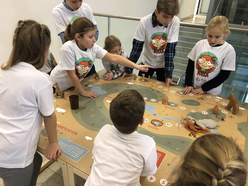 Dzieci ubrane w białe koszulki stoją wokół stołu, którego blat jest gra planszową (źródło: muzeumgniezno.pl)