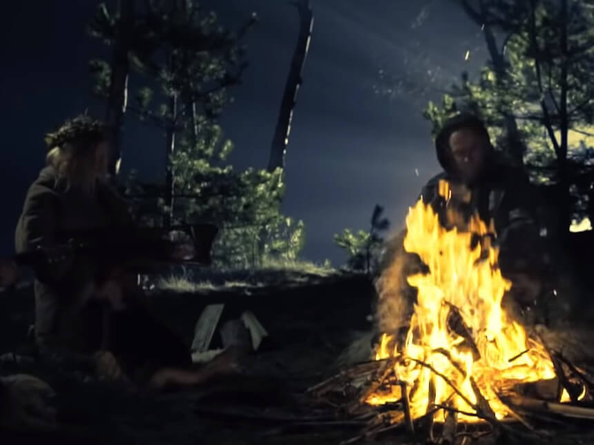 Noc w lesie. Przy ognisku siedzą - zakapturzony mężczyzna i kobieta z wiankiem na głowie. (źródło: youtube.com)