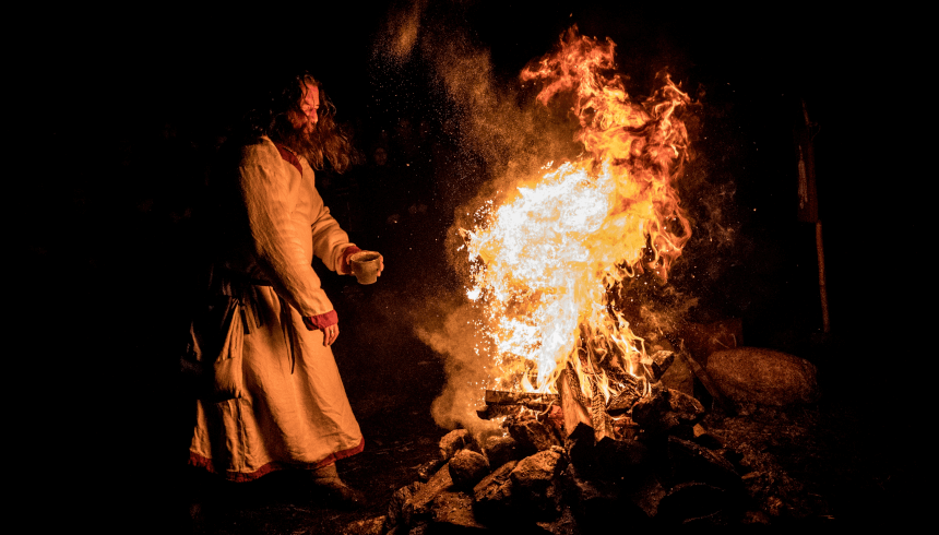 Rekonstrukcja obrzędu dziadów. Noc. Mężczyzna w słowiańskim stroju stoi przed wielkim ogniskiem. Ciało ma zwrócone w stronę ognia. W lewej ręce trzyma gliniane naczynie.