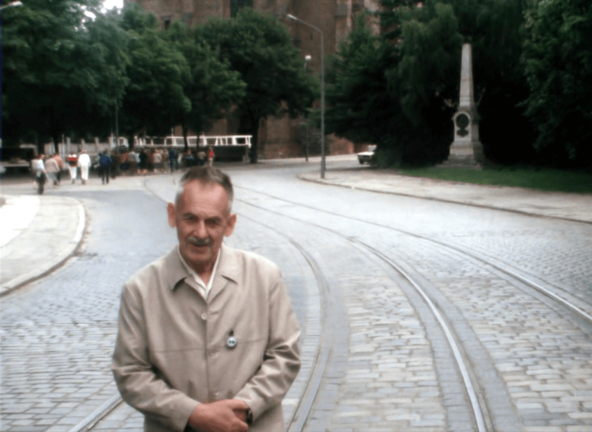 Starszy mężczyzna - przewodnik turystyczny, na tle Ostrowa Tumskiego w Poznaniu. Stoi na brukowanej drodze prowadzącej do poznańskiej katedry.