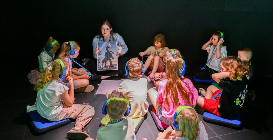 dzieci (w słuchawkach na uszach) siedzące wokół edukatorki trzymającej w rękach obrazek