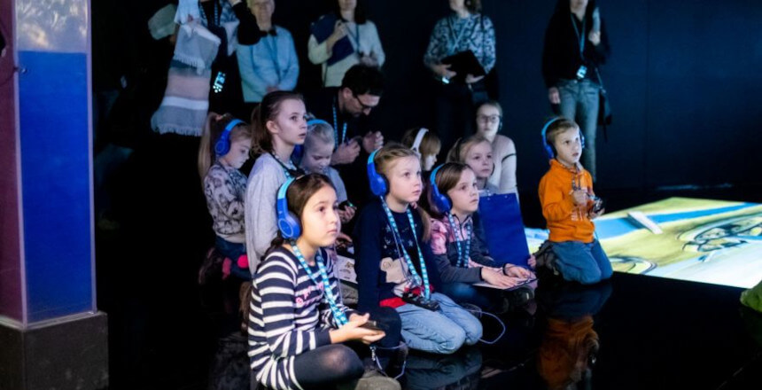 Grupka dzieci, wyposażonych w słuchawki i audioprzewodniki, zwiedza ekspozycję Bramy Poznania. 