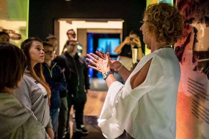 Kobieta - przewodnik, opowiada o ekspozycji muzeum korzystając z języka migowego. Przed nią młodzież.